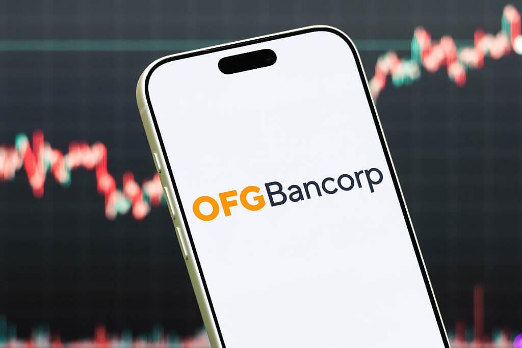 Celular mostrando el logo de OFG Bancorp con gráficas bursátiles de fondo en rojo y verde
Ilustración fotográfica de: Cheng Xin/Getty Images