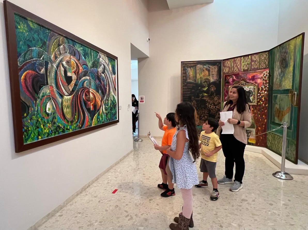 Grupo de niños observa pinturas en una galería de arte con guía sosteniendo papeles y señalando las obras.
Foto: Vía Facebook: Museo de Arte de Bayamón