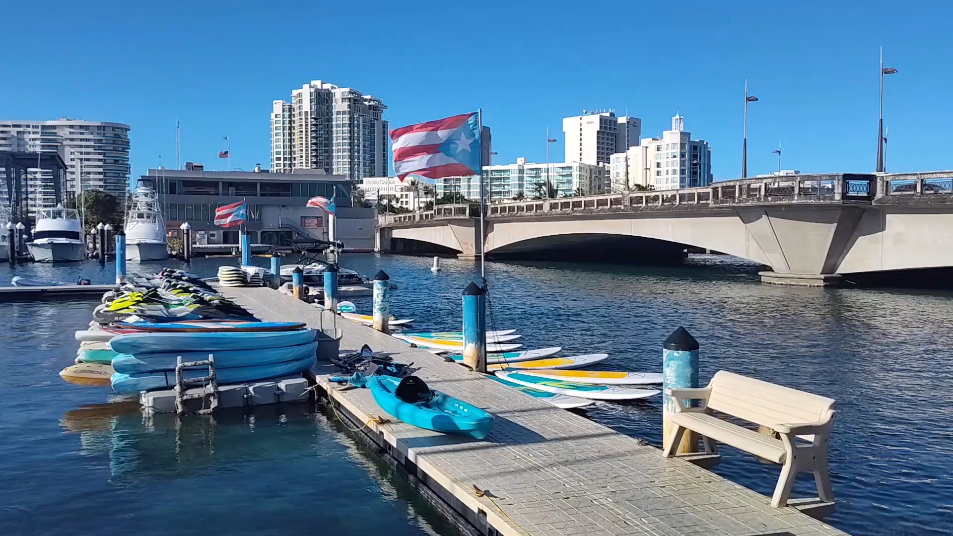 Muelle con kayaks y tablas en la Laguna del Condado, con la bandera de Puerto Rico ondeando frente al puente Dos Hermanos