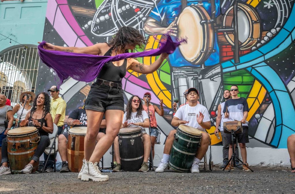 Bailarina de bomba con un pañuelo violeta frente a barrileros y un mural colorido durante un bombazo callejero